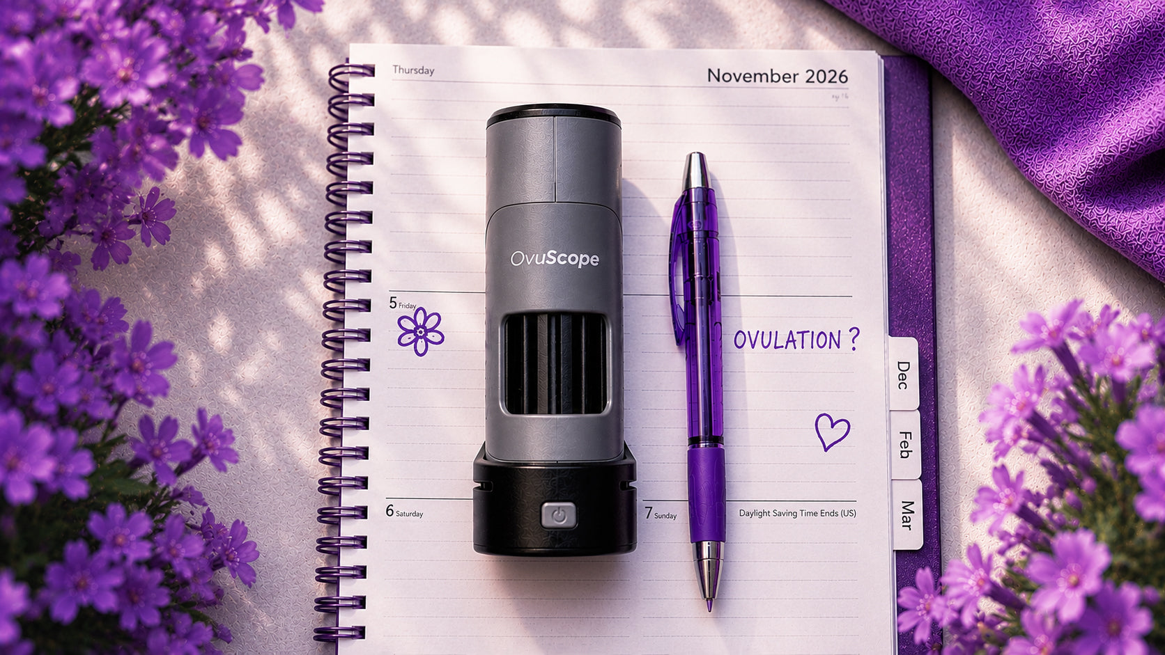 A compact cylindrical ovulation tracker device in gray and black sits on an open spiral notebook with a purple pen beside it. The scene is styled with purple flowers and a purple fabric corner, suggesting a lifestyle or flat-lay setup.